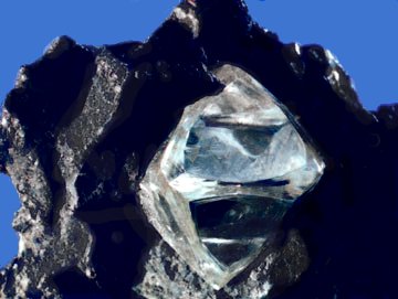 Clear, glassy diamond with an octahedral shape embedded in a black chunk of rock.