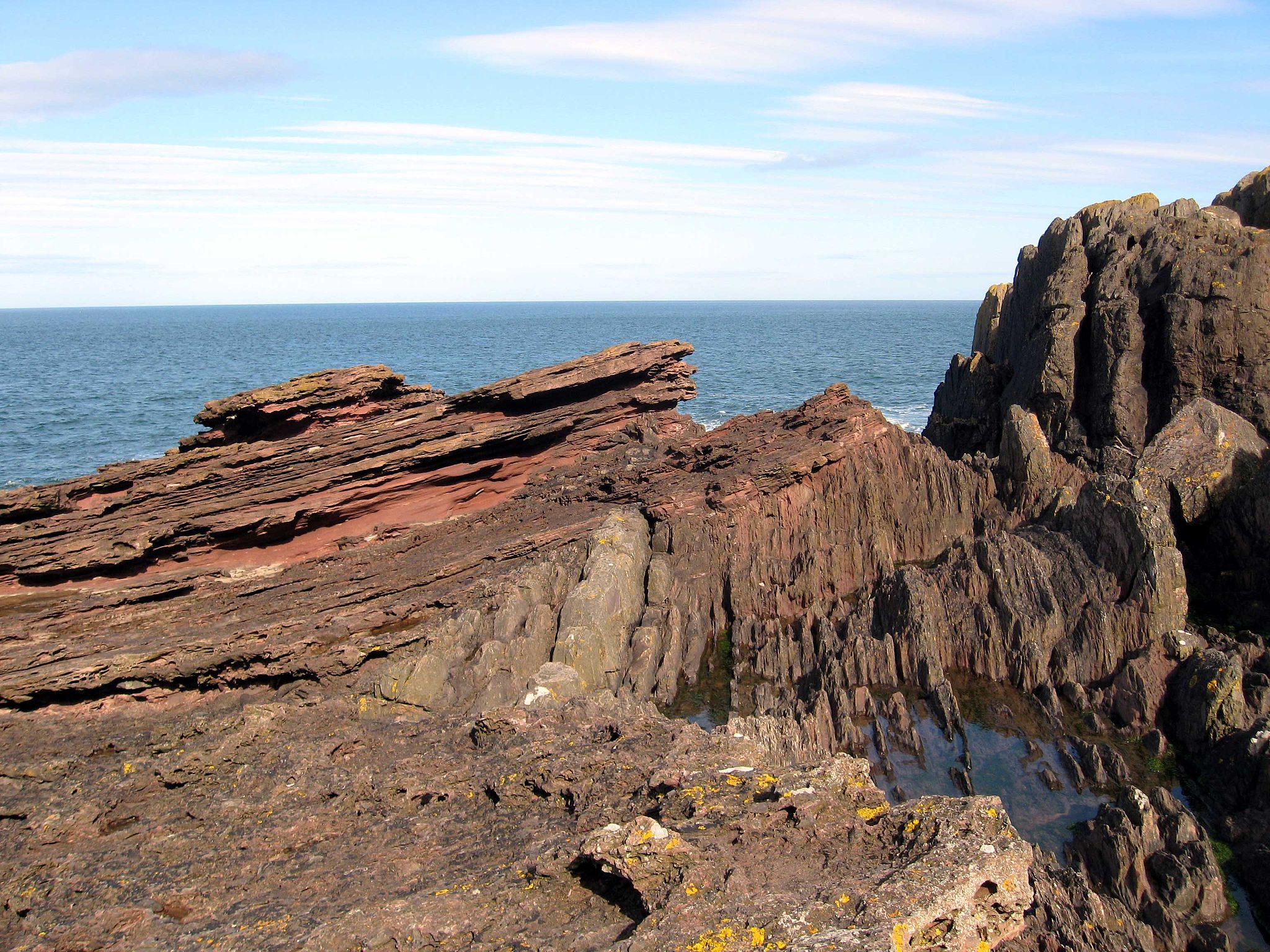 On the right hand side is a series of dark brown rock layers that are vertical. As you look toward the left, the vertical layers are capped by gently-dipping reddish brown sedimentary layers. The ocean is seen in the background.