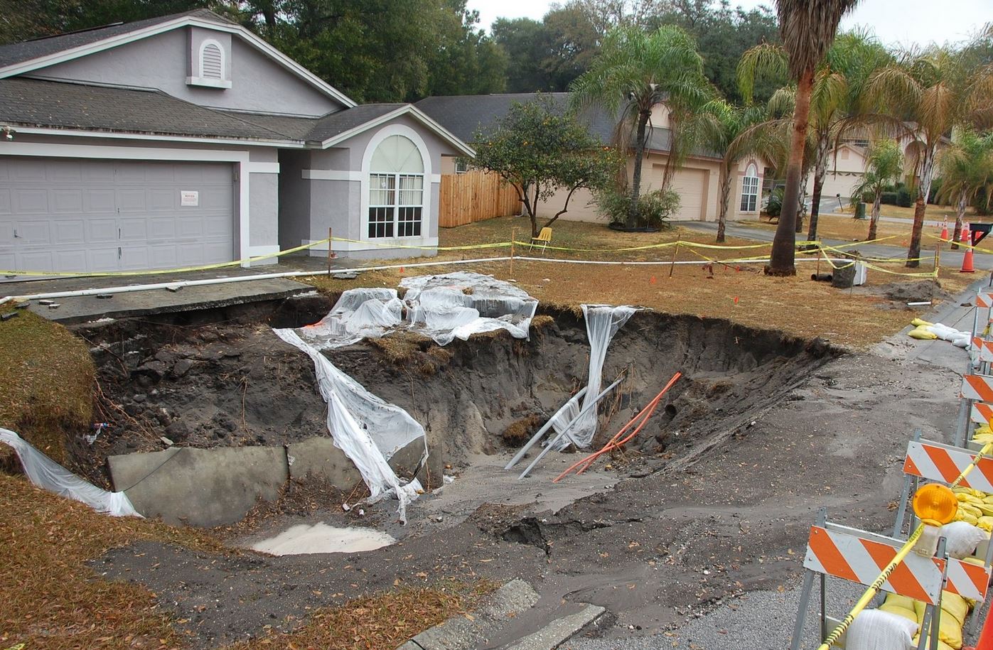 Dirt-lined sinkhole in front of a gray house; the sinkhole is cordoned off with caution tape.