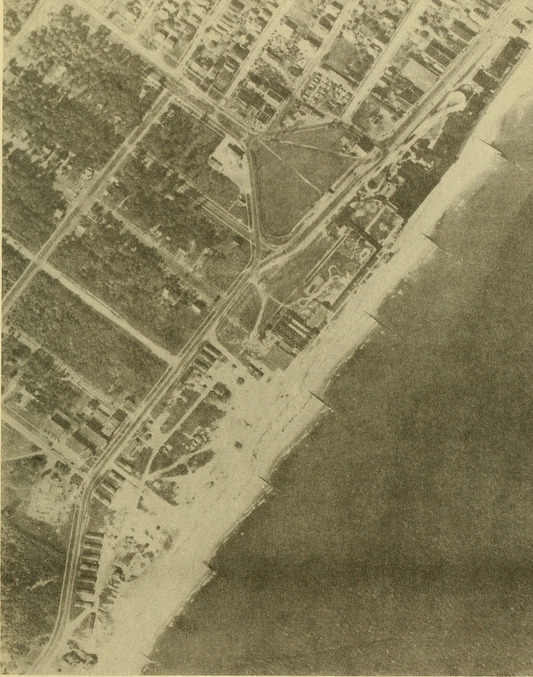 Black and white aerial photo of a series of parallel groins extending perpendicular outward from the shoreline with buildings visible on the mainland; sand is built up on beach on the left side of each groin and sand is eroded away from the beach on the right side of each groin.