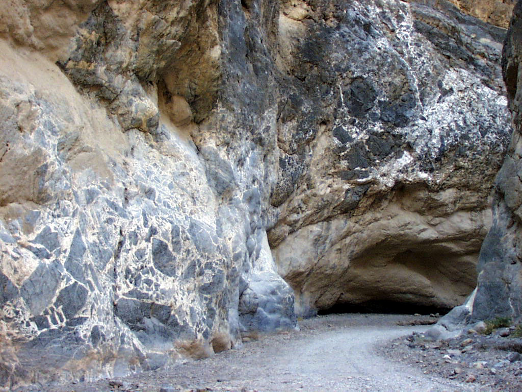 Cliff face alongside a curving gravel road; numerous large, angular dark gray boulder fragments are embedded throughout the white cliffside rock.