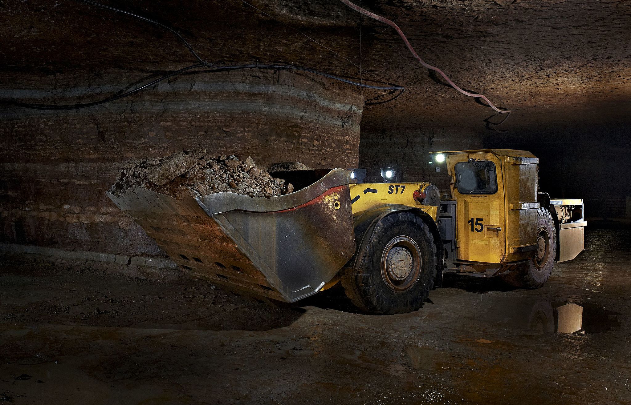 A photo in an underground mine with horizontal layering visible in the mine walls; there is also a large yellow truck with a bucket attached to its front containing brown rocky material.