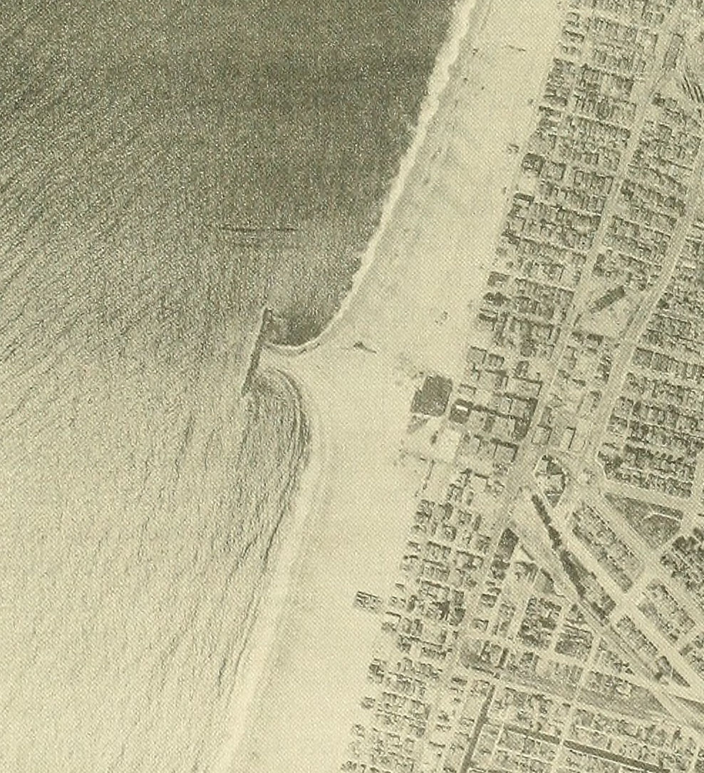 Black and white aerial photo of a breakwater that was built in shallow ocean water parallel to the shoreline but currently has sand built up from the beach all the way to the structure. Buildings are visible on the mainland.