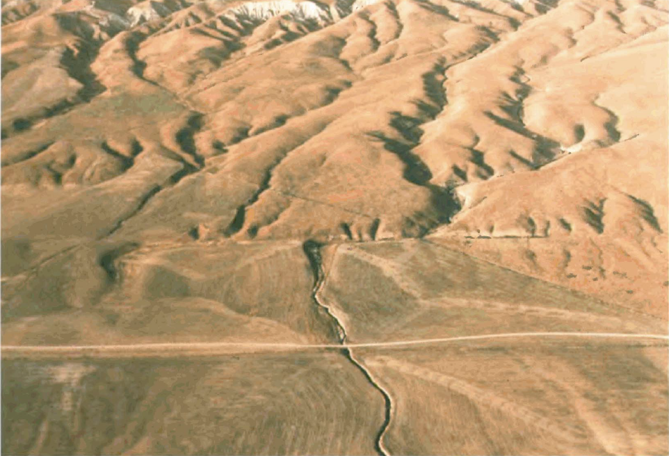 Aerial photo of a landscape in California. A dry creek flows from the northern mountainous part of the image, then takes a sharp right as viewed from the flow of water, then a sharp left, caused by the San Andreas Fault cutting roughly perpendicular to the creek. The fault can be seen about halfway down, trending left to right, as a change in the topography.