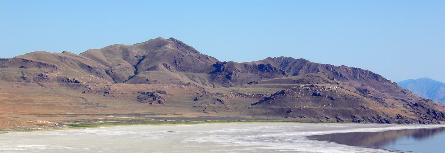 Panoramic view of erosional notches in a brown hillside with a white playa and part of a lake in the foreground.