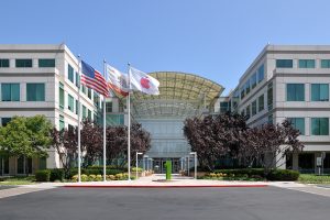 A photograph of Apple Headquarters in Cupertino, California. Two square buildings sit on the right and left of the photo, with an arched ceiling and entrance in the center. Trees sit in front of the entrance, with three flagpoles to the left of the entrance.