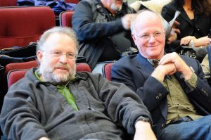 A close up photograph of Ben Cohen (right) and Jerry Greenfield (left) sitting in stadium style theater seats, with people sitting around them.