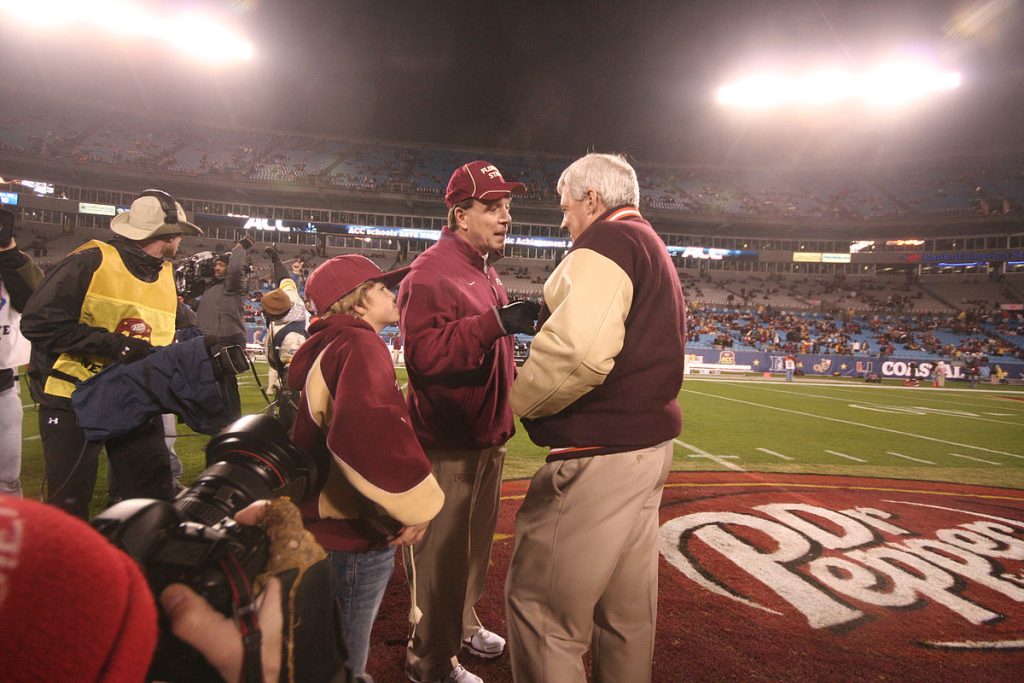 Frank Beamer stands to the right of Jimbo Fisher on a football field in a stadium with people in the stands at night.