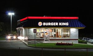 A photograph of a Burger King at night, as seen from a road. The building is lit up with lights on it, and a streetlight is in the background. A car is at the drive thru window with its headlights on.