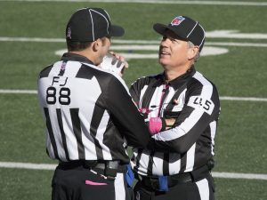 A photograph of two referees talking on a football field. Both are wearing black and white striped shirts and black hats. One is standing with their back to the camera, and the other is facing the other referee toward the camera.