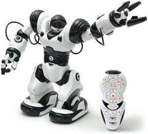 A photograph of a Robosapien, similar to the one pictured in figure 14.1, on a white background. The robot is standing, looking to the right, and holding its arm up toward the right corner. Beside the robot is a black and white remote, smaller than the Robosapien, sitting upright.