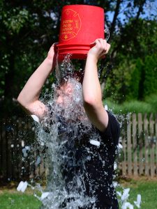 A photograph of a person standing outside, holding a red bucket upside down above their head. Out of the bucket, water and ice is falling on the person, obscuring their face and upper torso.