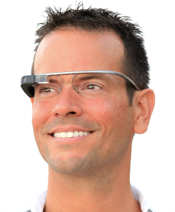 A photograph of a person wearing Google Glass, a pair of silver glasses with a small black camera on the right side. The person is looking off into the distance to the left in front of a white background.