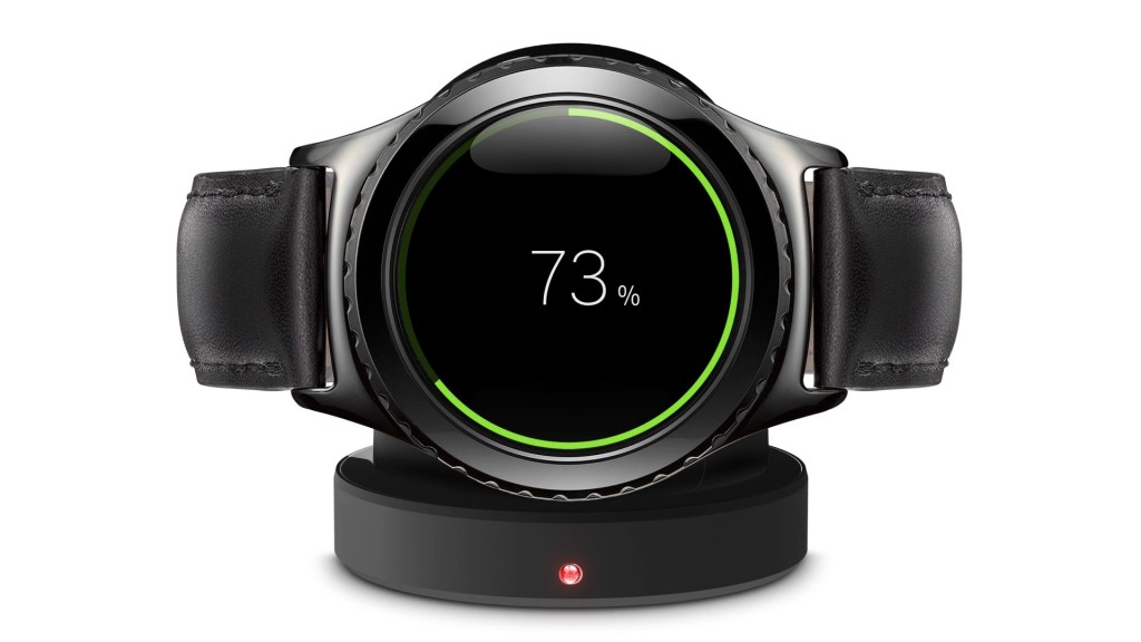 A photograph of a Samsung smart watch, sitting on its charger on a white background. The watch has a round face, and the face and band are black. A green ring around the face shows the charging percentage. The Percentage is displayed on the face, 73%. The charger has a red light in the middle.