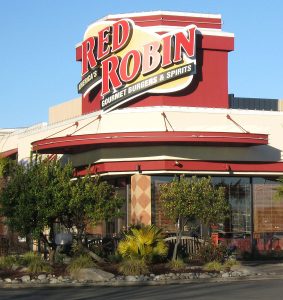 A photograph of the outside of a Red Robin restaurant, as seen from the road. The Red Robin sign sits on top of the building, and shrubs sit in front of the building.