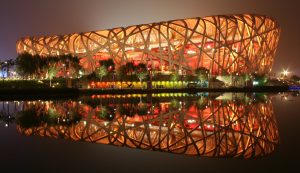 A photograph of the Beijing National Stadium at night, taken from across water. The National Stadium is lit up by yellow lights from within. A reflection of the stadium appears on the water.