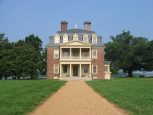 A photograph of the Shirley Plantation, shown from the front with a path leading up to the house. The house is three stories tall, with five windows on each story. A porch on the front of the house is two stories high. Trees are lined up on either side of the house, with a blue sky in the background.