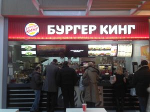 Photograph of the inside of a Burger King restaurant, with people standing in line to order. The Burger King store name is in Russian.