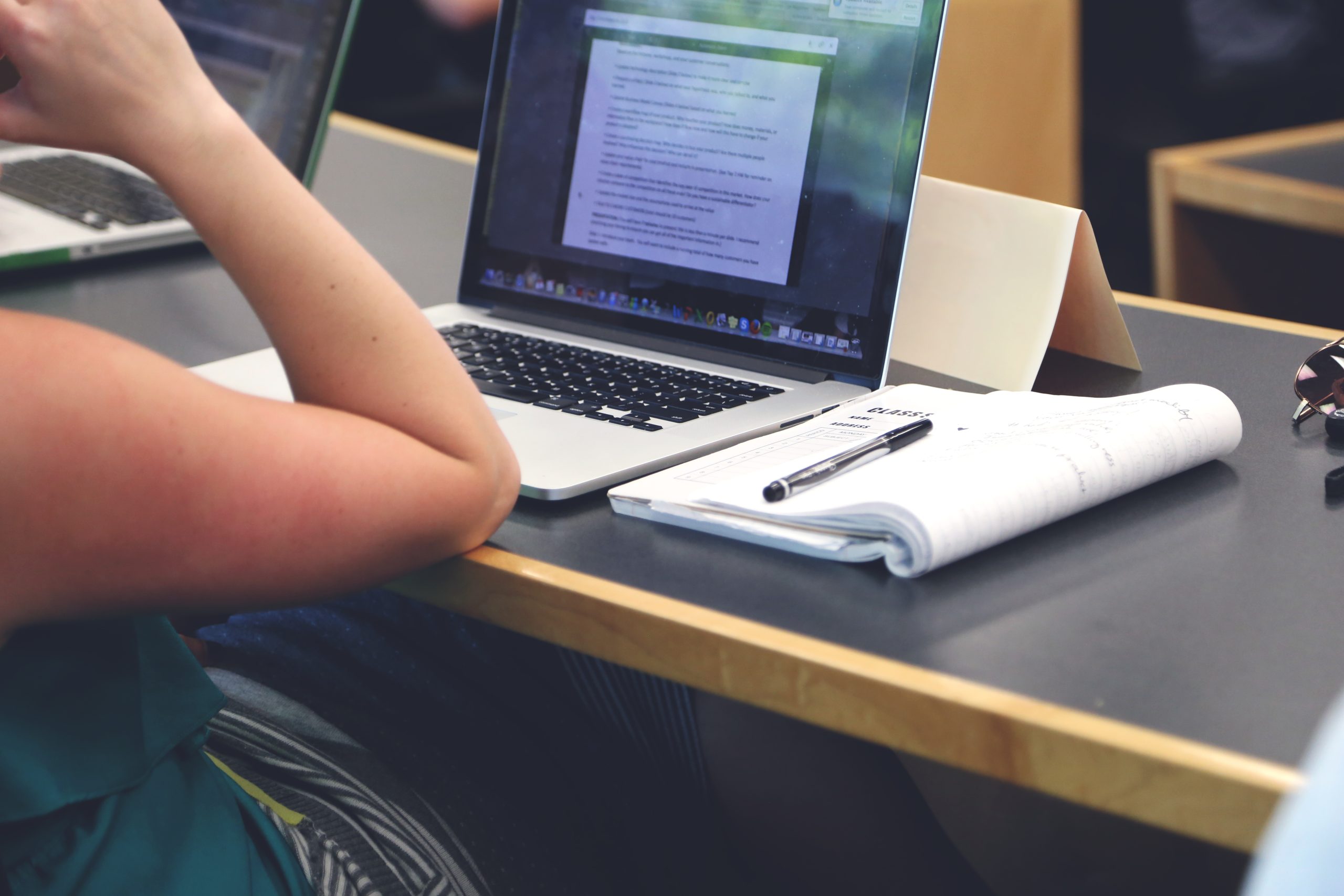 A photograph of a person sitting at a table with their laptop open to a window filled with text. A notebook is open beside the laptop with a pen on top.