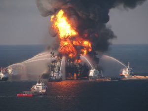 A photograph of an oil rig engulfed in flames with dark smoke billowing upward. Five boats surround the oil rig, spraying water on it to stop the fire.